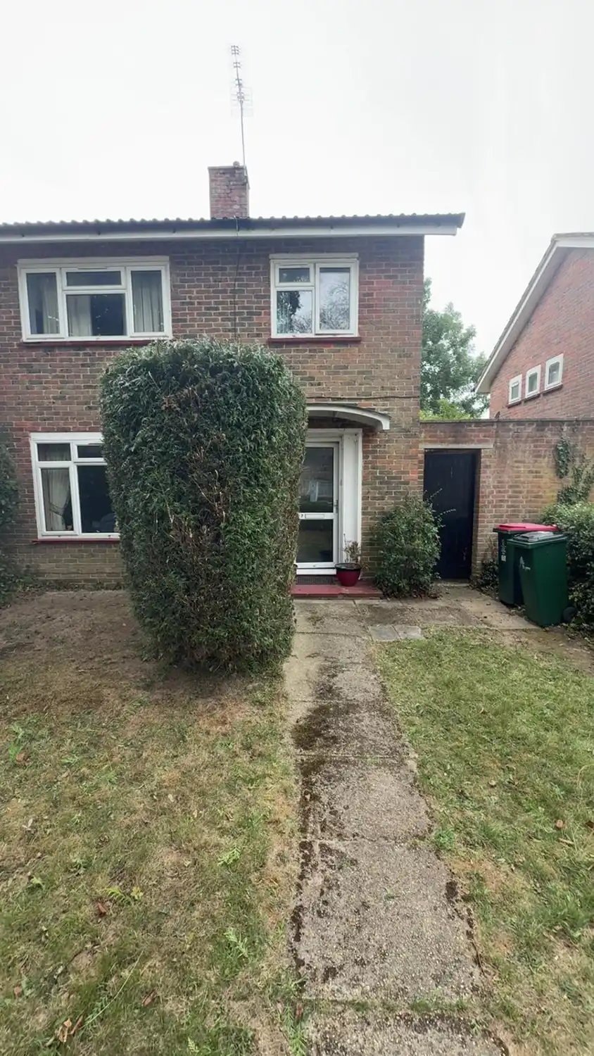 Front view of a brick two-story house with three windows and a front door partially obscured by a large, rounded shrub on the left side of a concrete walkway leading from a patchy lawn. A smaller shrub and two green and red garbage bins are visible to the right of the entrance, next to a black gate in a brick wall.